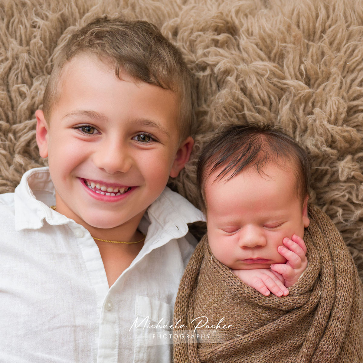 Älterer Bruder liegt lachend neben seinem schlafenden neugeborenen Geschwisterchen, das in braune Stricktücher gewickelt ist.